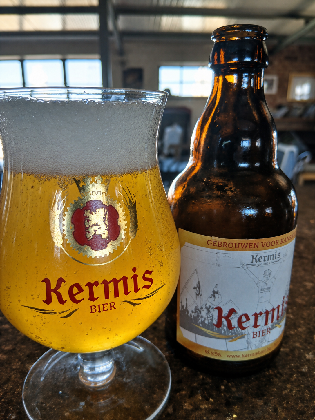 A glass of golden Kermis Blond beside its bottle in a Belgian café setting